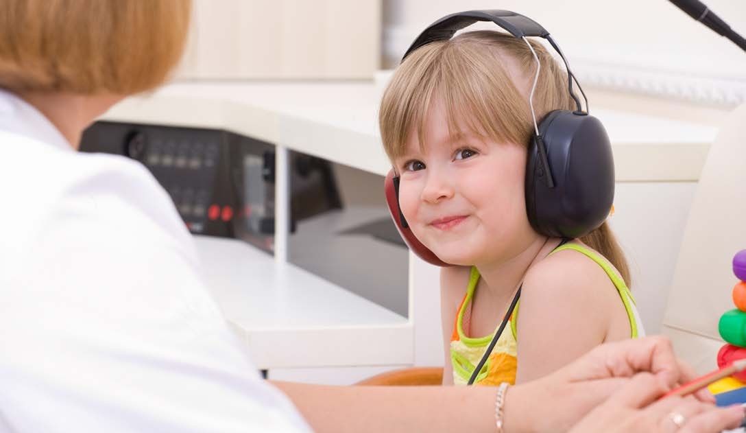 Young girl wearing headphones at the audiologist’s office, smiling at the doctor