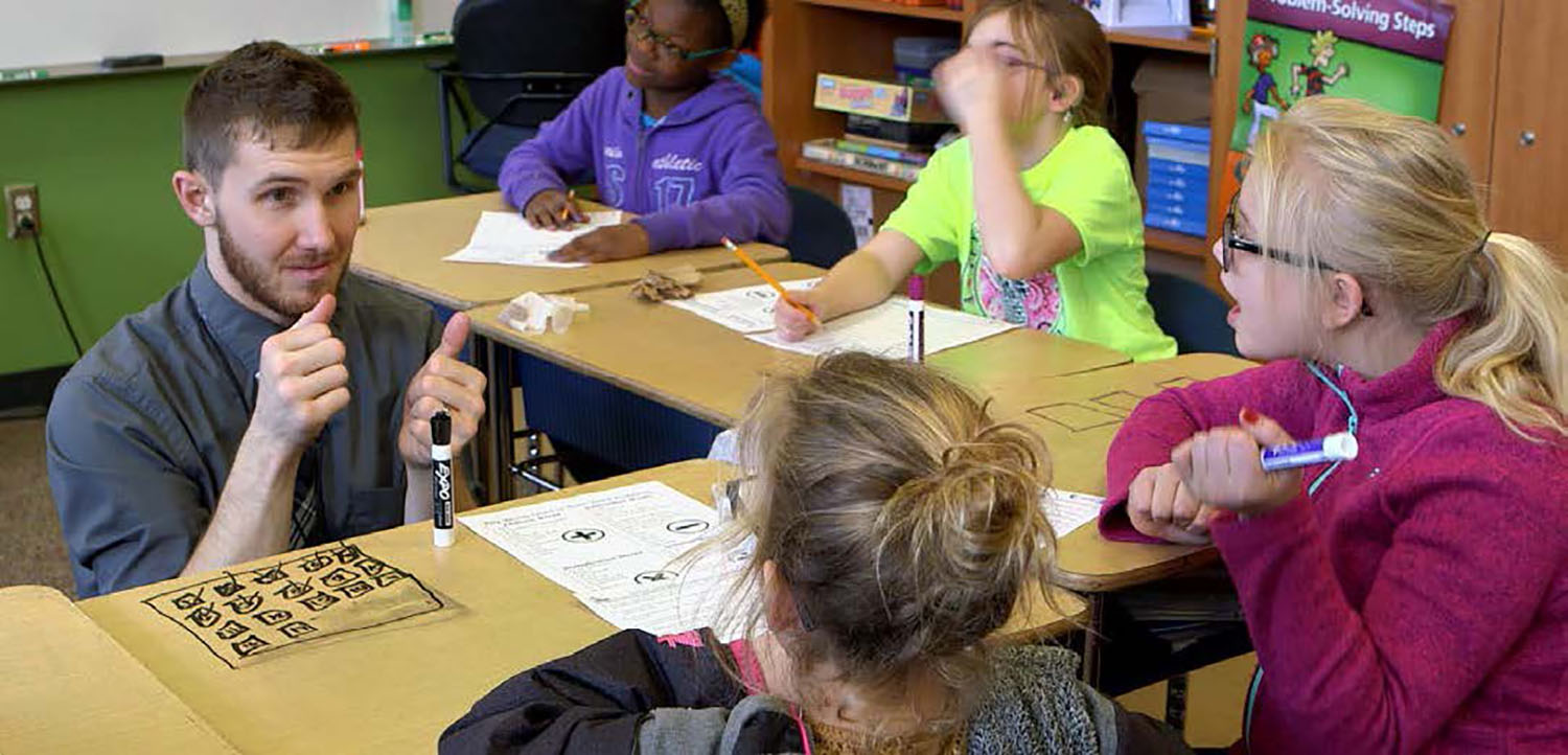 U-shaped seating in a classroom with students working at their desks and a teacher of the deaf in the middle explaining the topic in sign language to two of the students.