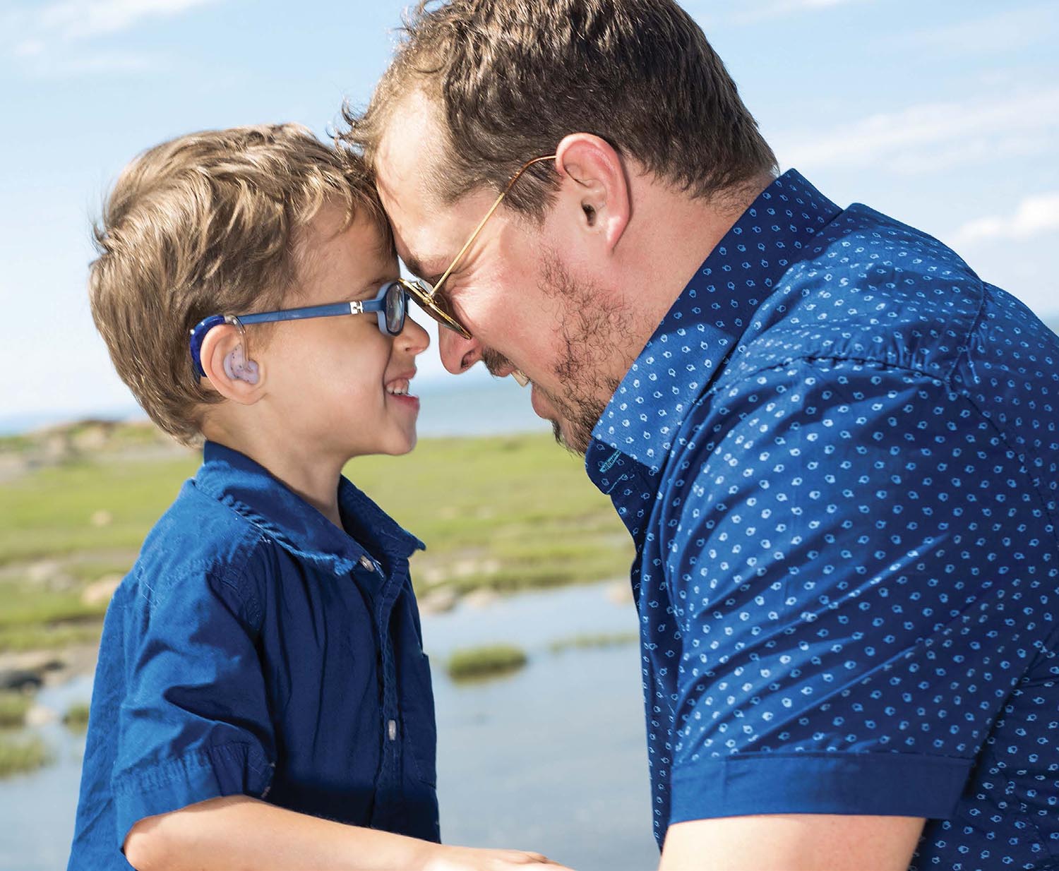 A young boy wearing a hearing aid and his dad, both wearing glasses, standing facing each other with their foreheads touching and big smiles.