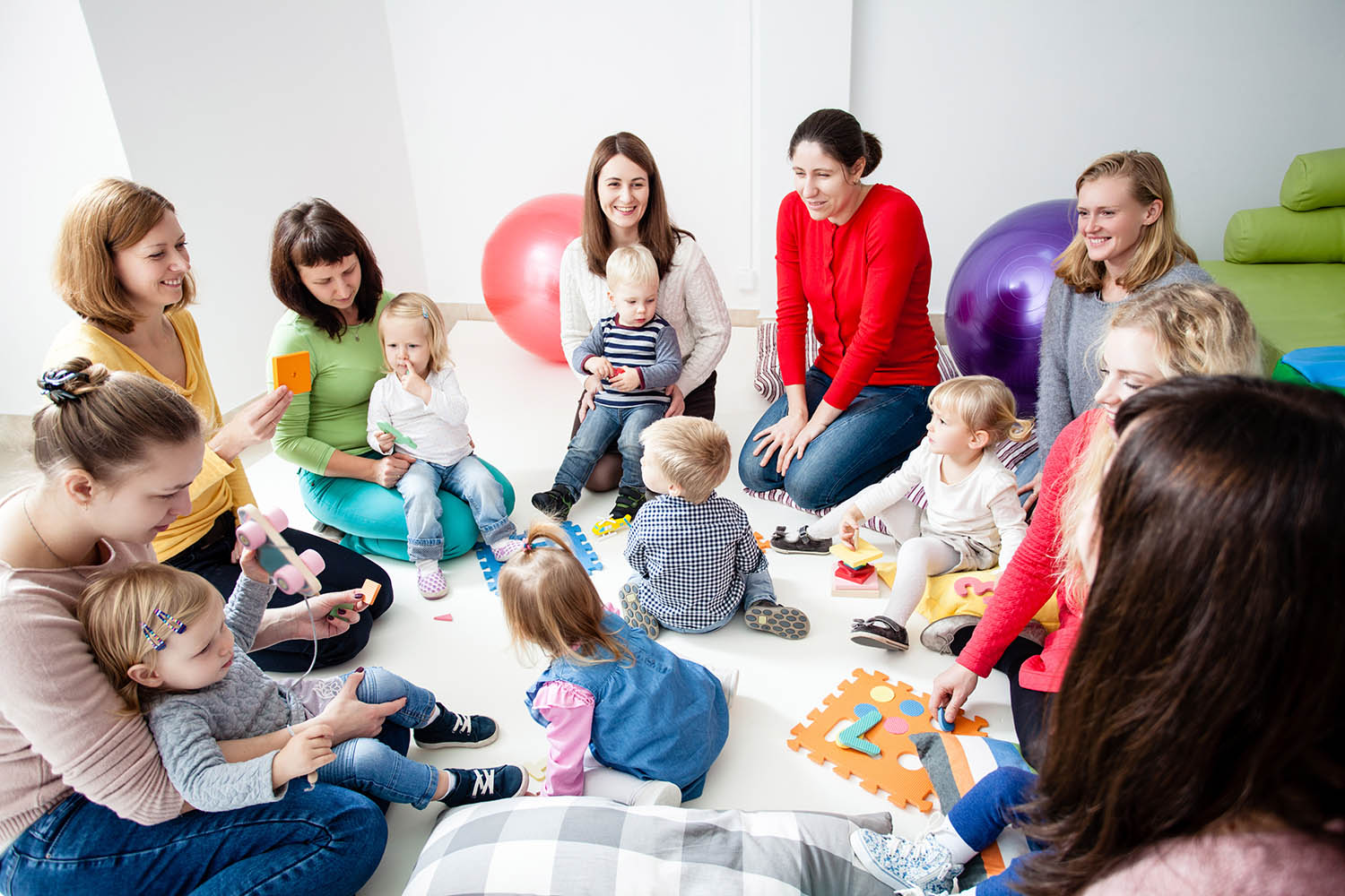 A group of parents sitting in a circle with children in their laps and in the center of the circle. Various toys are around the room.