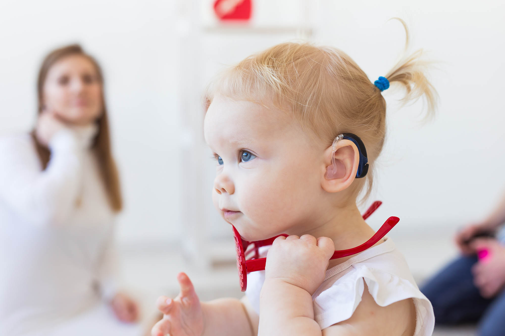 Smiling toddler wearing a hearing aid with parents in the background.