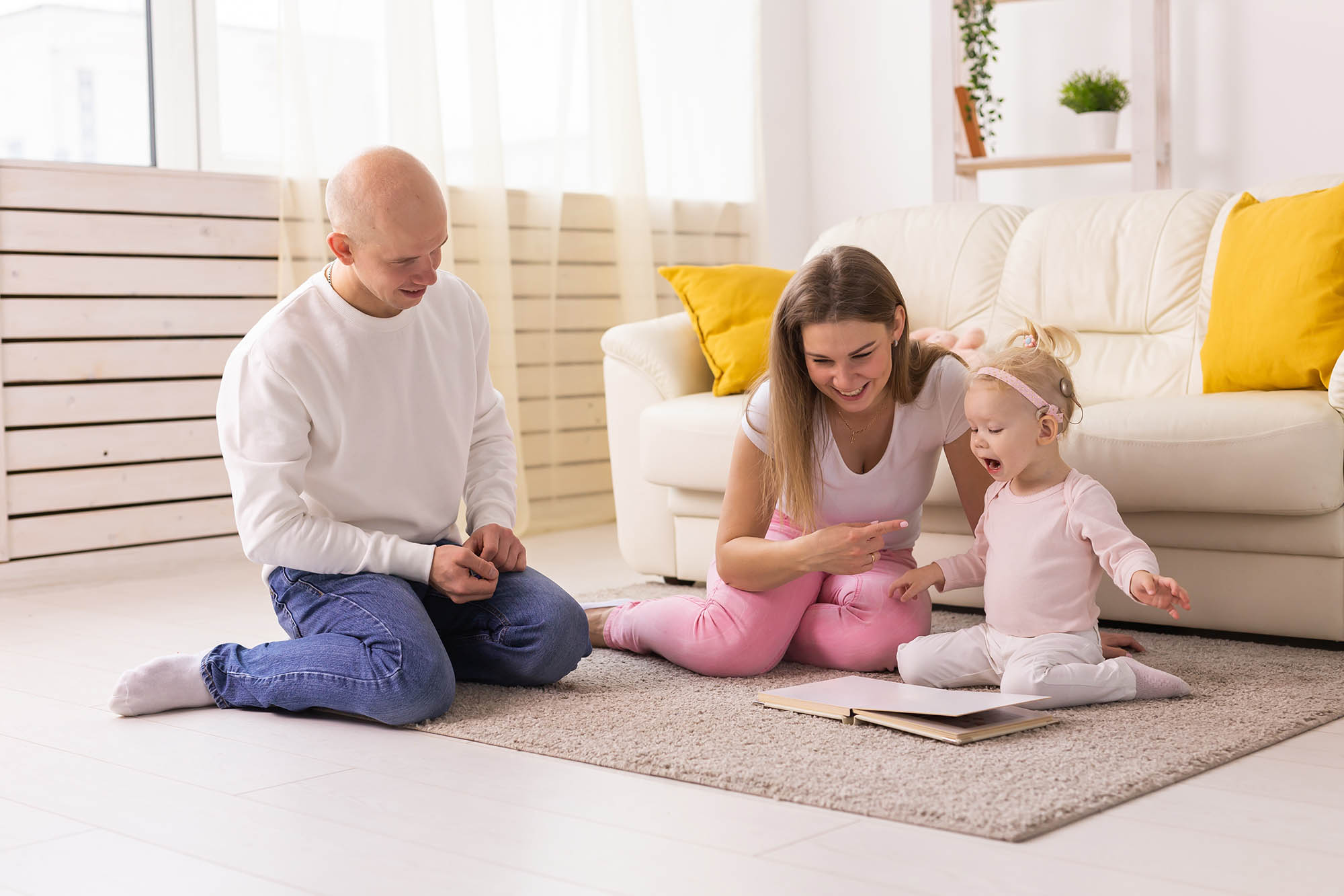A child sitting upright on the floor looking at a book with her parents who are also sitting on the floor
